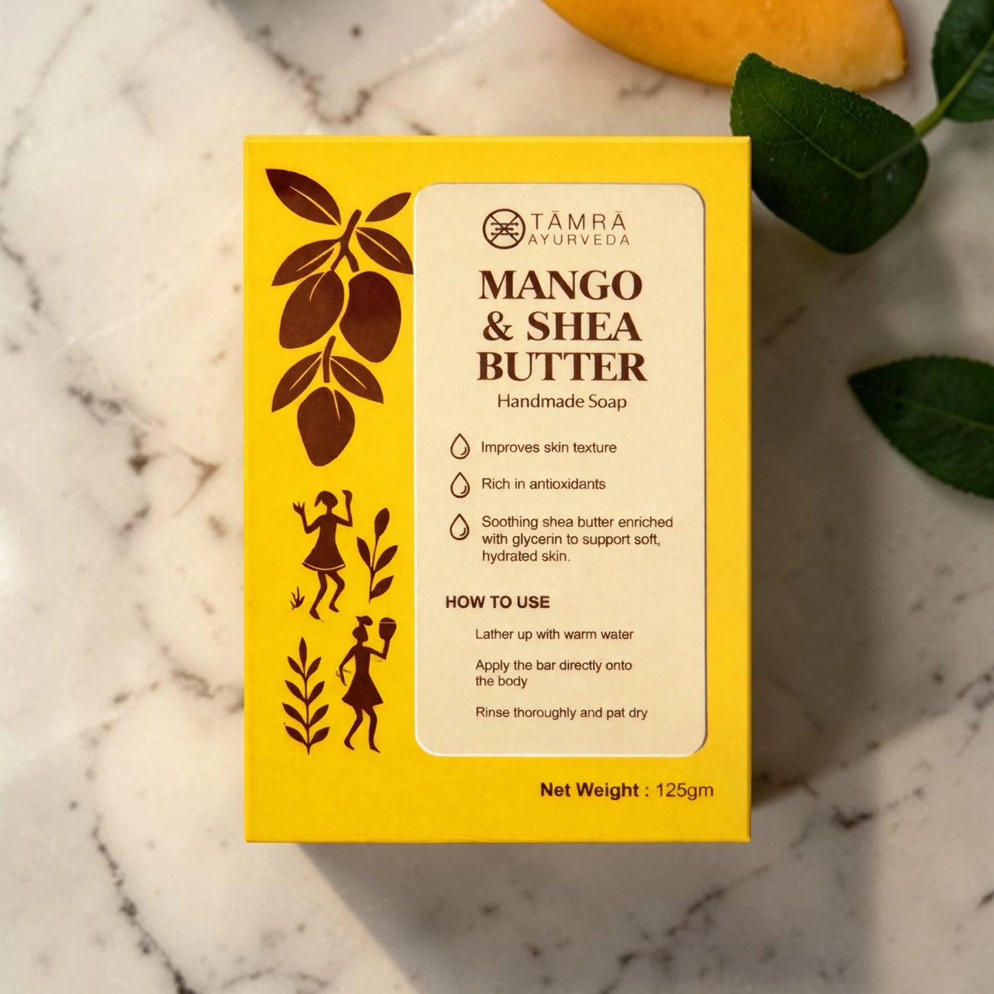 Yellow box of Tamra Naturals Mango & Shea Butter soap on a marble surface with leaves.