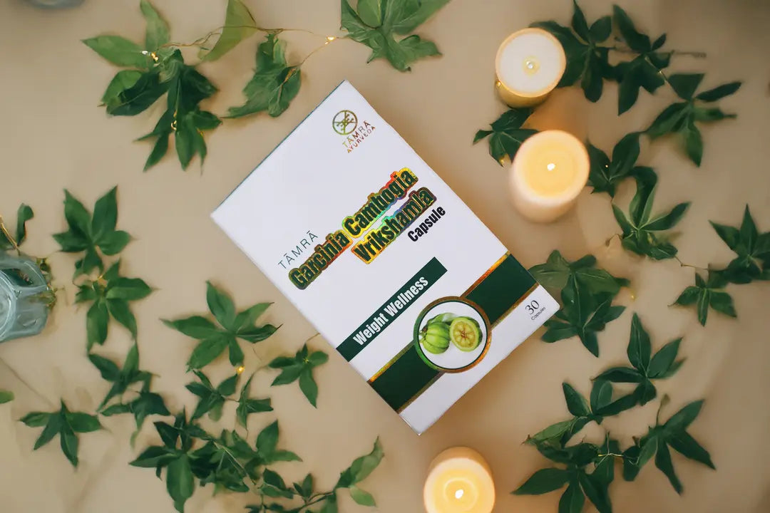 Box of 'Tamra Garcinia Cambogia Vrikshamla' capsules surrounded by green leaves and candles on a beige surface