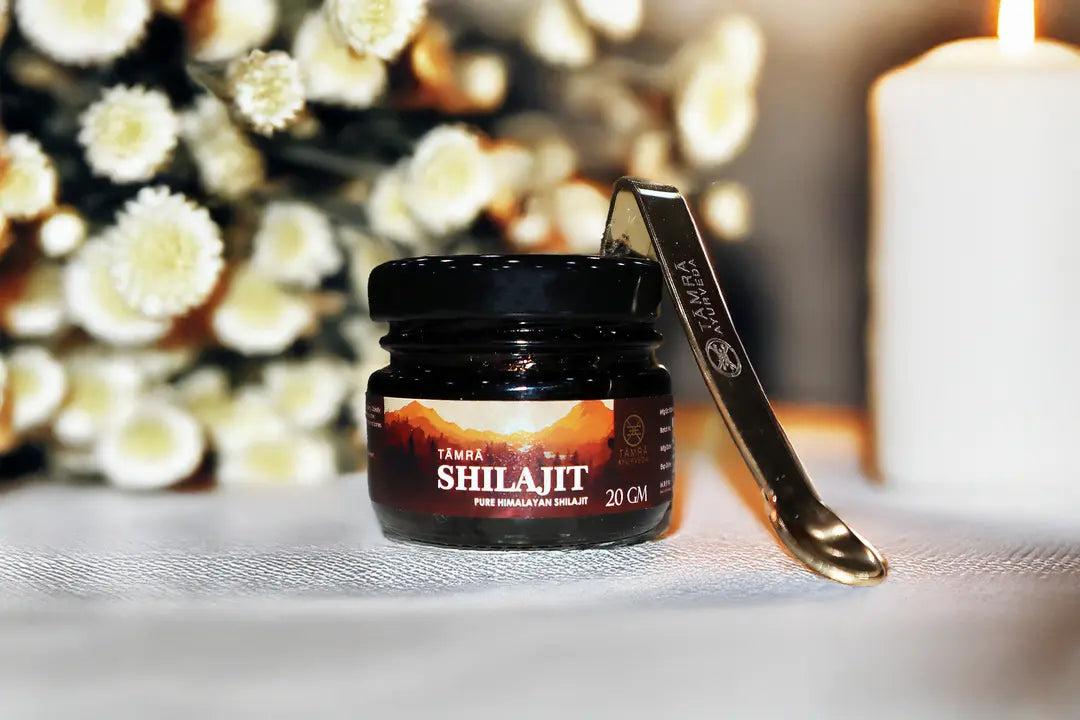 Jar of Tamra Ayurveda Shilajit with a spoon on a decorative surface with flowers and a candle.