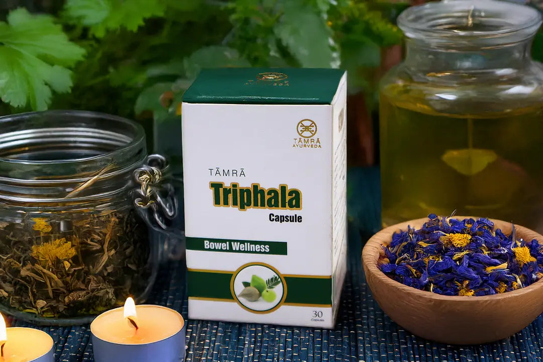 Box of Tamra Triphala Capsules with jars and a bowl of dried flowers on a textured surface.