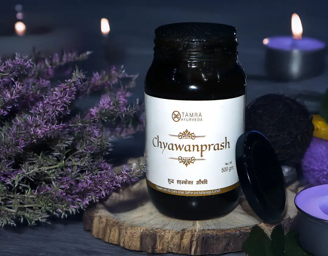 Jar of Chyawanprash with Tamra Ayurveda label on a wooden platform with purple flowers and candles in the background.