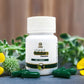 White bottle labeled 'Yamra Goldaura Capsule' with green capsules on a wooden surface with yellow flowers and green leaves.